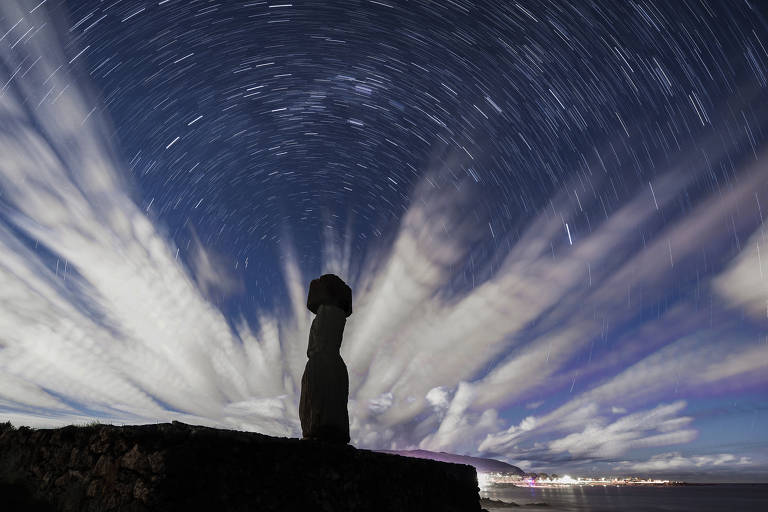 Um único moai aparece como uma sombra contra o céu noturno, iluminado por estrelas e por nuvens. Ao fundo, bem discretamente, é possível ver luzes da cidade à distância