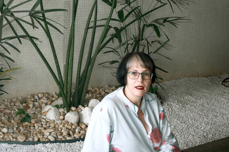 A retratada posa em frente a um ornamento de plantas, que estão contra uma parede branca. Ela olha para a câmera, e veste uma camisa branca. Só é possível vê-la da cintura para cima