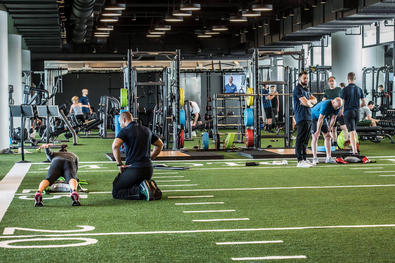 A academia do clube tem chão forrado em carpete que imita um campo de futebol americano. O espaço é amplo, e há vários equipamentos de musculação espalhados no espaço, com pessoas usando quase todos