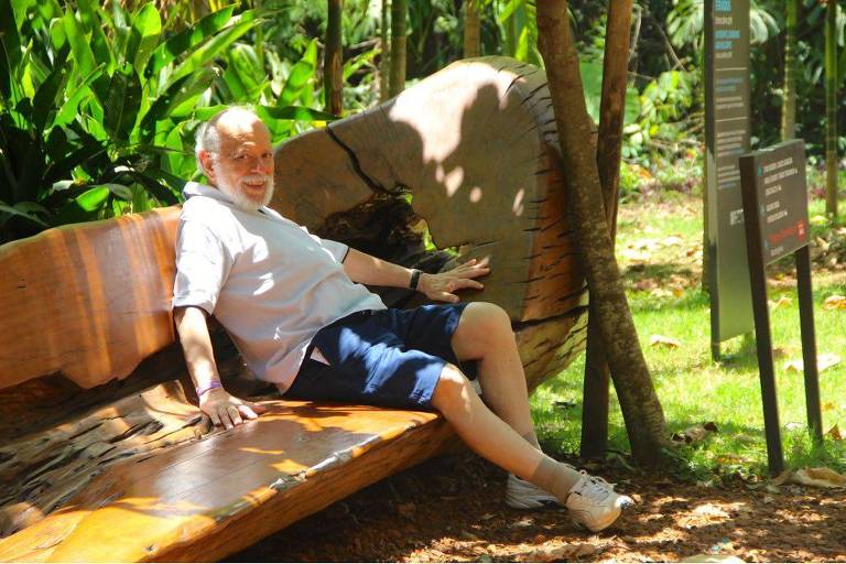 Foto mostra o arquiteto Luiz Carlos Costa sentado de camisa e bermuda, sorrindo, em banco de madeira no parque do centro Inhotim, perto de BH