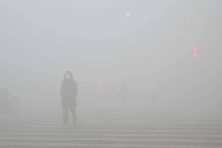 Pessoa, caminha em meio a smog (nuvem tóxica), em um dia poluído em Weifang, uma província de Shandong, em dezembro de 2016. No meio da poluição, a imagem da pessoa fica ligeiramente apagada. Ao fundo, é possível ver luzes verdes e vermelhas de semáforos.