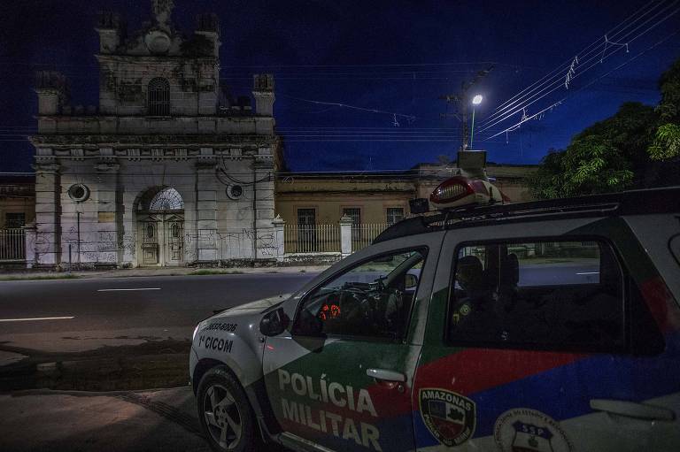 Viatura da Polícia Militar do Amazonas estacionada em frente à Cadeia Pública Raimundo Vidal Pessoa, em Manaus, que se vê ao fundo, de noite