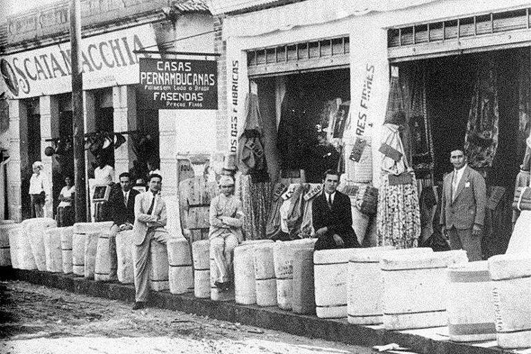 Fachada de uma das primeiras lojas da Pernambucanas, com produtos dispostos na calçada e pessoas olhando a câmera