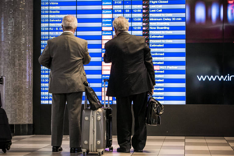Dois homens de terno, de costas, olham para painel eletrônico com informações sobre voos em aeroporto
