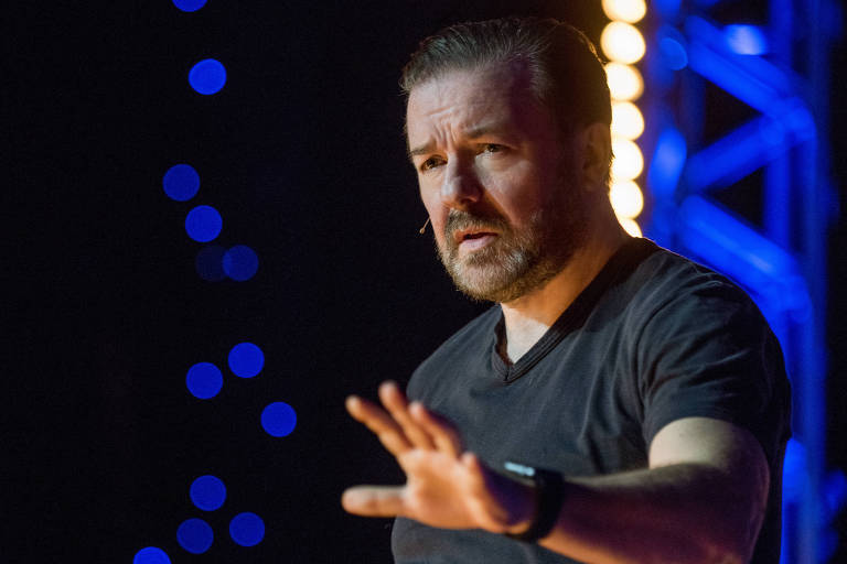 Foto mostra o comediante Ricky Gervais fazendo gesto de atenção com a mão, sobre o palco com luzes azuis