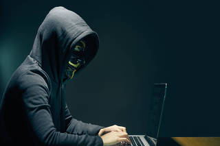 Side view of person wearing hooded top and Guy Fawkes face mask using laptop