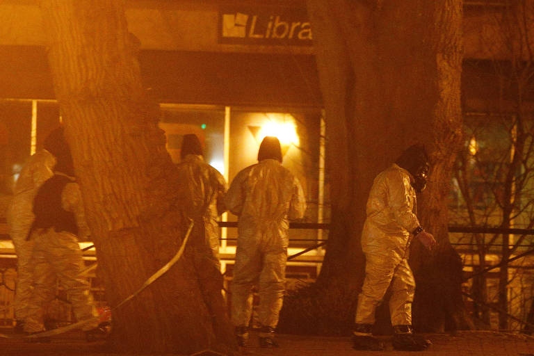 Durante uma noite, quatro peritos são vistos à distância de pé, com roupas de proteção contra agentes químicos, ao lado de duas árvores; a planta da esquerda está com uma fita de isolamento do local; ao fundo, uma loja com uma lâmpada amarela acesa