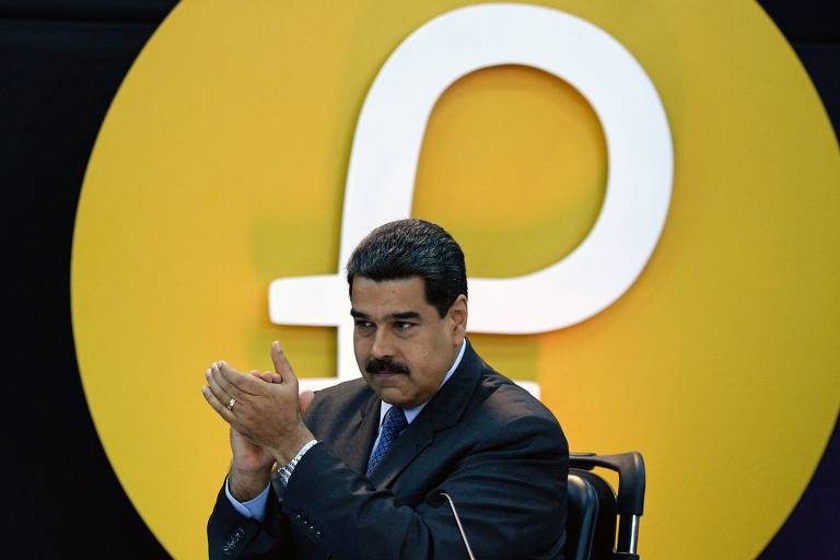 Sentado, o ditador venezuelano, Nicolás Maduro, aplaude em um evento no Palácio de Miraflores; ao fundo, o símbolo do petro, em um círculo amarelo sobre um fundo preto