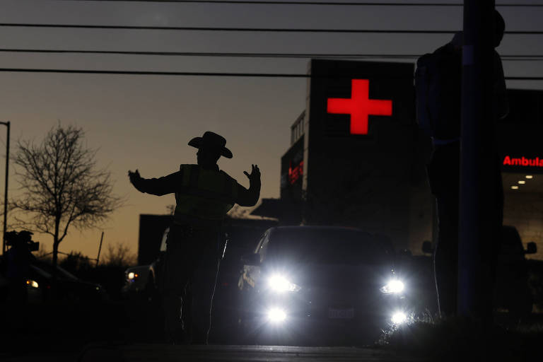 De chapéu de caubói, policial aparece redirecionando o tráfego em meio ao fim de tarde em Austin, no Texas; homem aparece sob penumbra e, ao seu lado direito, destacam-se os faróis de um carro e a cruz acesa de um hospital