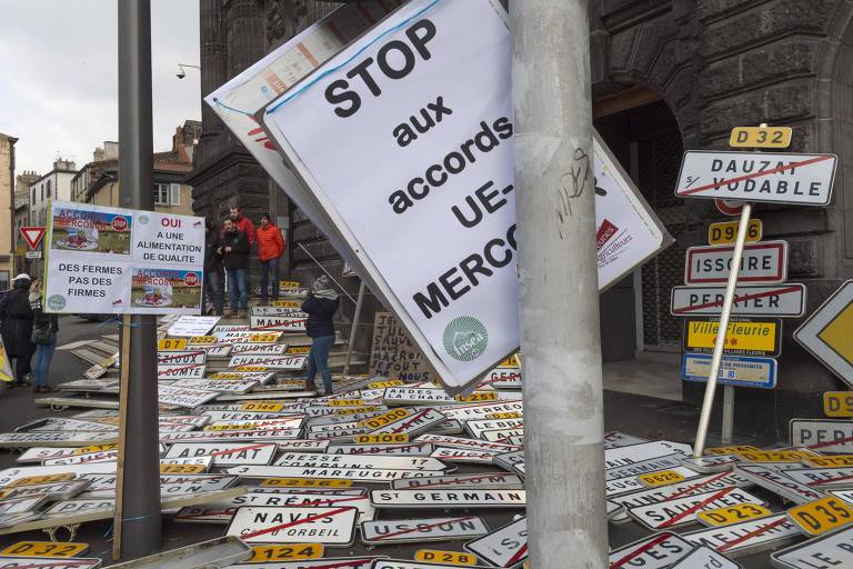 Placas de diferentes municípios franceses são vistas empilhadas no chão, enquanto em dois postes são pendurados cartazes contra o acordo de livre-comércio entre a União Europeia e o Mercosul