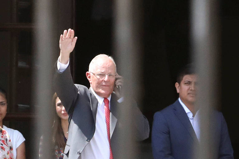 Pedro Pablo Kuczynski acena com a mão esquerda enquanto fala ao celular com a mão direita ao sair do palácio de governo em Lima; imediatamente atrás, aparecem assessores; imagem foi feita de fora da grade do palácio, que aparece na foto