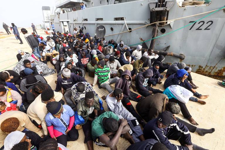 De cabeça baixa, grupo de imigrantes africanos espera sentado em um cais ao lado de uma embarcação da Marinha da Líbia no porto de Zawiyah