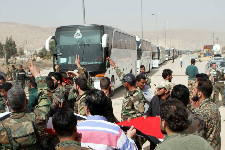 De roupas verdes e camufladas, combatentes rebeldes levantam a mão ante policiais que escoltam uma fila de ônibus rodoviários usados para deslocá-los de Harasta, em Ghouta Oriental, para o nordeste da Síria