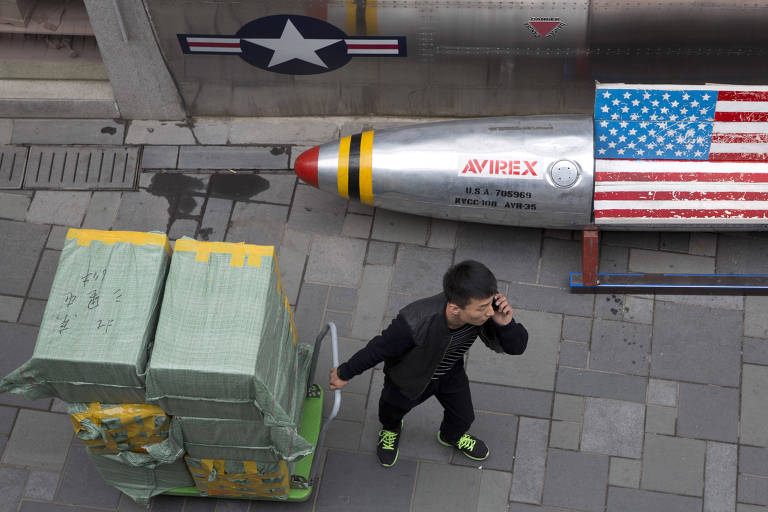 Com carrinho com caixas verdes empilhadas, homem chinês de preto fala ao celular ao passar pela calçada de uma loja de produtos americanos em Pequim; à direita, um banco com formato de foguete com assento com formato da bandeira dos EUA
