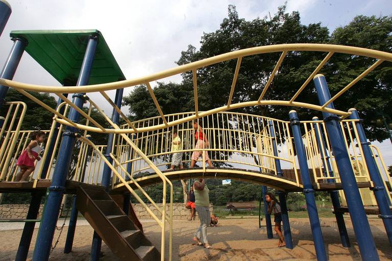 Quatro crianças brincam em um brinquedo do playground de um parque. O brinquedo tem uma ponte de madeira com estruturas de metal de proteção e uma grade para as crianças se pendurarem