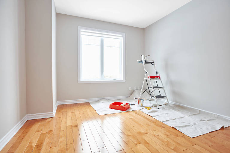 A foto mostra um cmodo vazio de um apartamento. O cho  revestido de madeira e as paredes so pintadas de branco. H uma janela ao fundo, e em frente, uma escada mvel