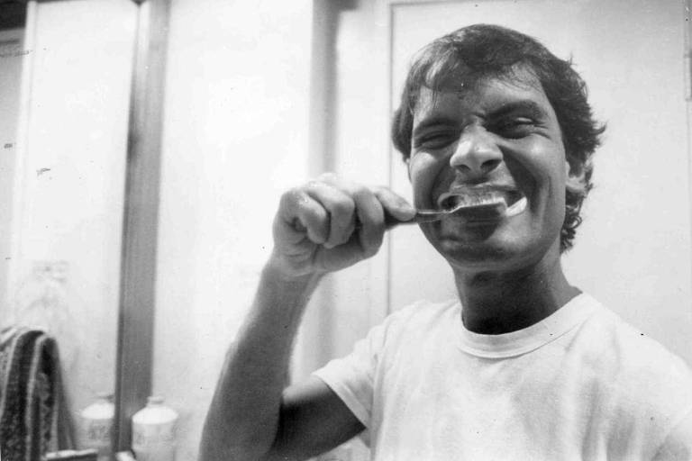 O cantor Cazuza escova os dentes. Ele sorri para a câmera com a escova em contato com os dentes e a boca cheia de pasta