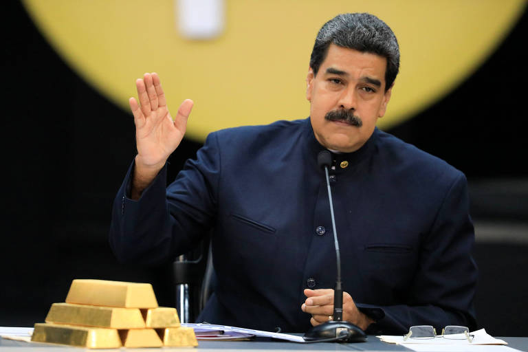 De camisa social preta, o ditador da Venezuela, Nicolás Maduro, levanta a palma da mão esquerda enquanto fala durante evento econômico; na mesa à qual está sentado, aparecem à direita papéis presos por um óculos de leitura, um microfone ao centro e imitações de barras de ouro à esquerda; o cenário é preto com um círculo amarelo com o símbolo do petro, a moeda virtual chavista, aparecendo pela metade