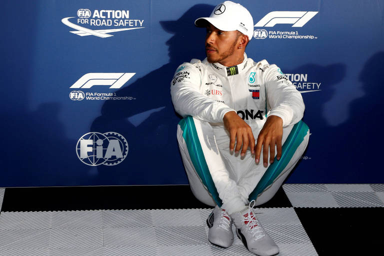 Lewis Hamilton descansa sentado com os pés cruzados e apoiando as costas em um backdrop azul