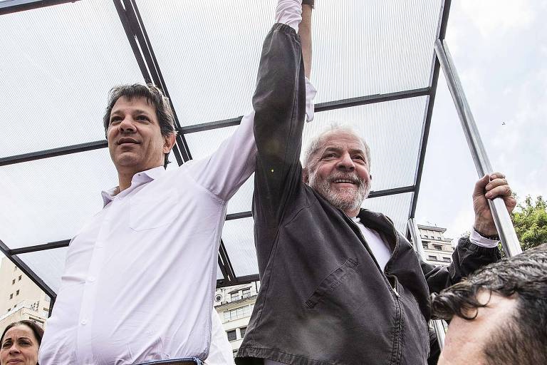 Fernando Haddad e Lula de mão dadas e erguidas sobre carro de som fazem campanha