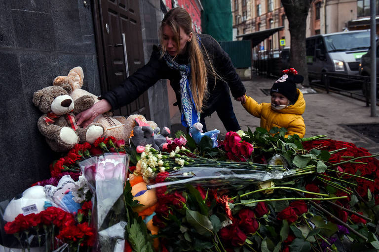 Mulher, segurando uma criança pela mão, coloca um ursinho de pelúcia em meio a flores em memorial pelas vítimas do incêndio