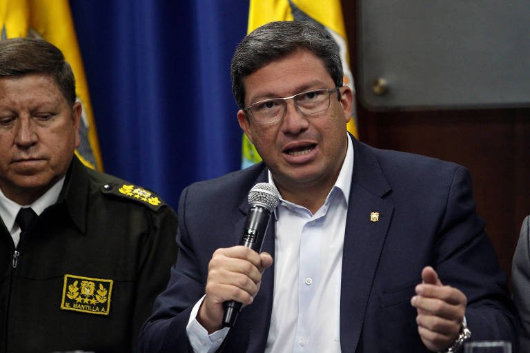O ministro equatoriano do Interior, César Navas, segura microfone ao lado de um oficial militar durante entrevista coletiva em uma sala do ministério em Quito; ao fundo, uma bandeira equatoriana pendurada em um mastro