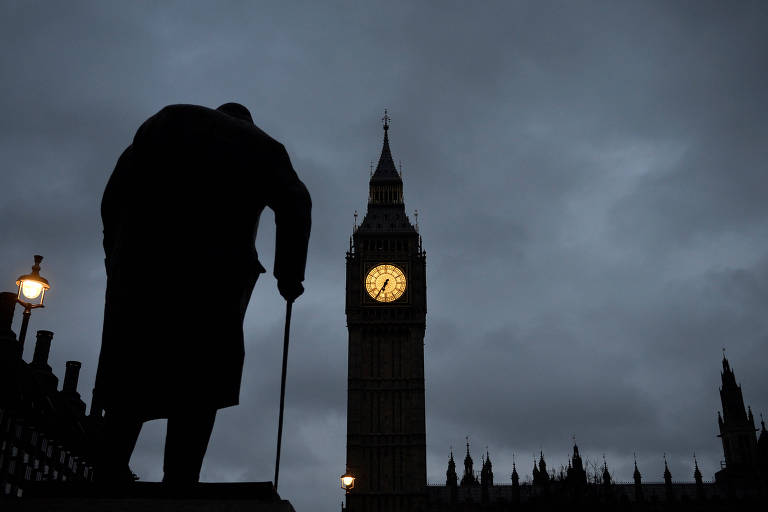 Estátua de Winston Churchill aparece sombreada e de costas, com o Big Ben e a sede do Parlamento ao fundo; céu nublado em fim de tarde ressalta as luzes amarelas do Big Ben e das lâmpadas em volta