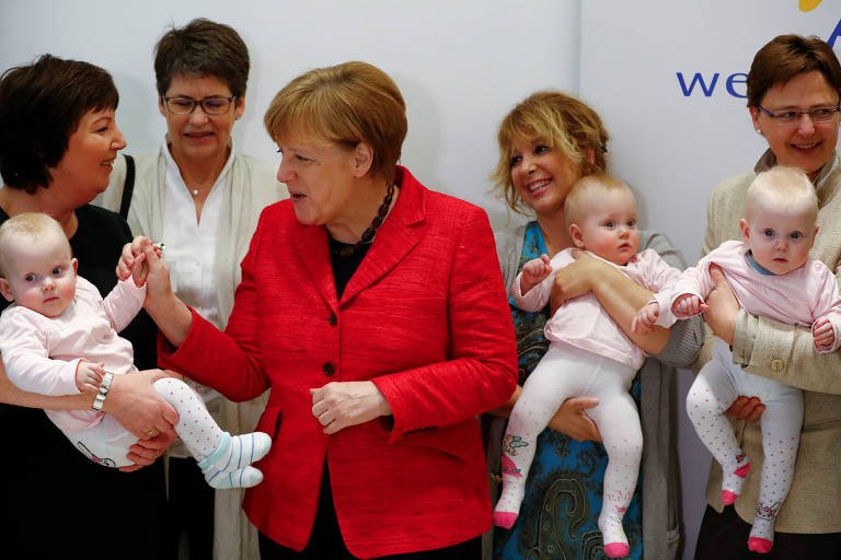 De terninho vermelho, a chanceler alemã, Angela Merkel, acaricia mão de bebê que é segurada por uma mulher de preto; outra mulher observa as duas no meio da imagem e outras duas pessoas aparecem à direita carregando as gêmeas da primeira criança