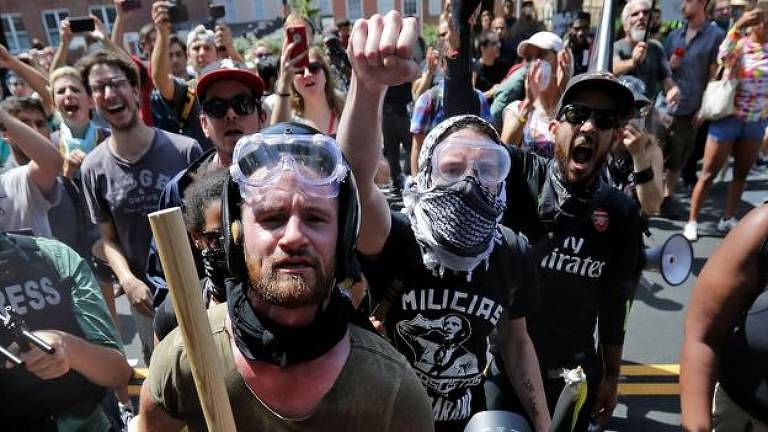 Grupo de manifestantes antifascistas gritam palavras de ordem durante manifestação em Charlottesville; à frente um homem com um lenço no pescoço e um pau na mão e imediatamente atrás outro, com um lenço quadriculado envolvendo o rosto e óculos de proteção, levanta o punho fechado