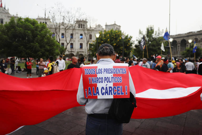 Homem carrega nas costas cartaz com as cores da bandeira peruana, vermelho e branco, em frente a um bandeirão peruano durante protesto em uma praça de Lima
