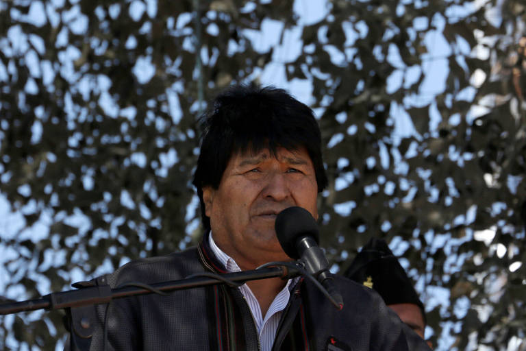 Evo Morales usa casaco preto com camisa branca e está diante de um microfone; ao fundo, folhas de uma árvore que lhe faz sombra
