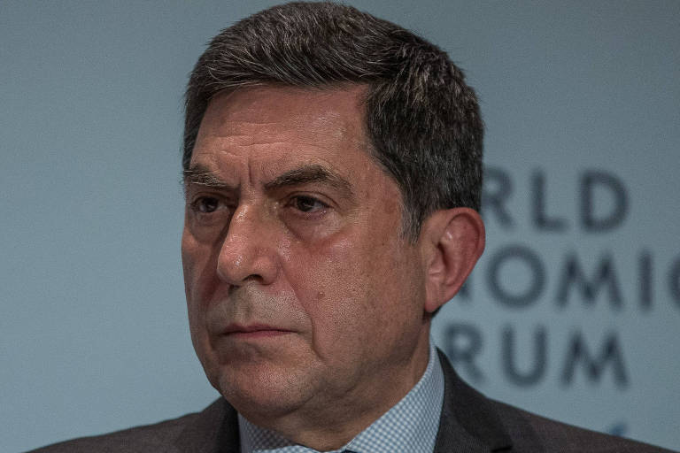 Luiz Carlos Trabuco é um homem branco com cabelos pretos e grisalhos. Ele veste terno cinza, com camisa e gravata azul. Ao fundo, há um painel do World Economic Forum.