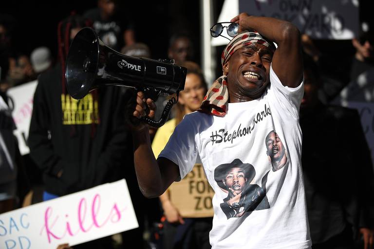 Com uma camiseta com uma foto do irmão, Stevante Clark leva o braço direito ao rosto enquanto chora ao falar do irmão em um protesto; com sua mão esquerda segura um megafone; atrás, manifestantes carregam cartazes