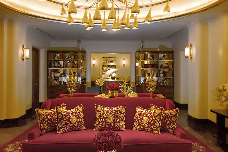 Mesa de centro com vaso de flores vermelhas. Atrás dele, um sofá vermelho com almofadas vermelhas e douradas. Ao fundo, prateleiras