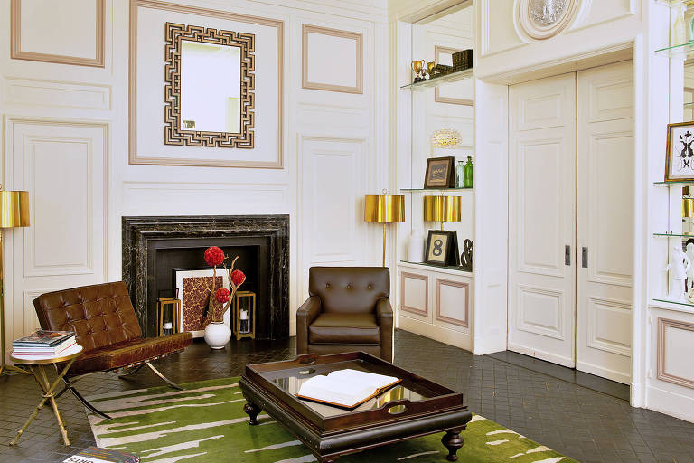 Sala de paredes brancas com tapete verde, mesa de centro e duas poltronas marrons ao fundo.