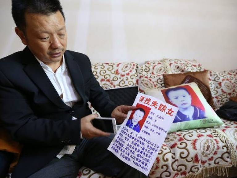 Wang Mingqing mostra cartaz com foto da filha; à direita, uma almofada com foto da menina repousa no sofá em que ele está sentado