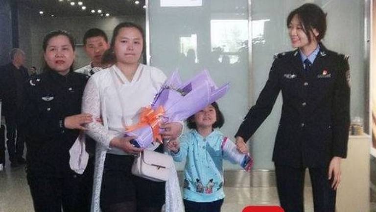 Kang Ying aparece com buquê de flores na mão ao lado de dois policiais em um dos saguões do aeroporto de Chengdu