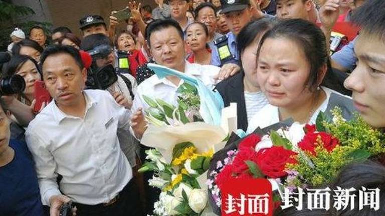 Com rosto emocionado, Kang Ying aparece com três buquês de flores nas mãos; três homens a acompanham: um à direita e dois à esquerda; atrás, jornalistas a observam