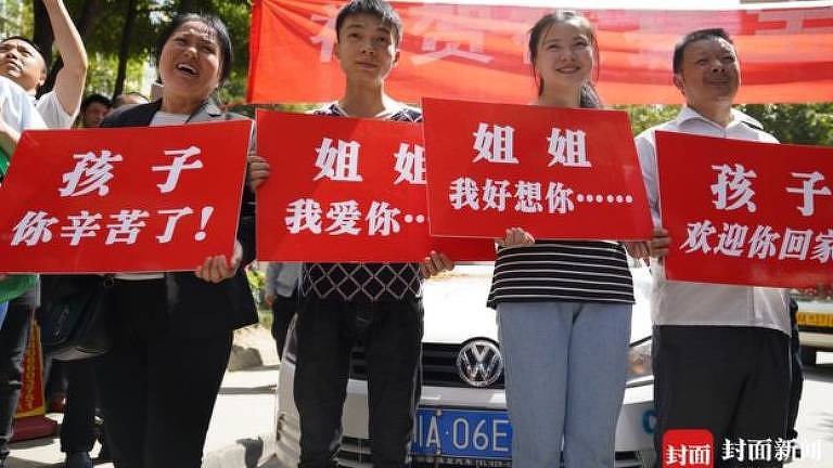 Familiares de Kang Ying carregam cartazes vermelhos de boas-vindas; ao fundo um carro estacionado e uma faixa vermelha mais alta