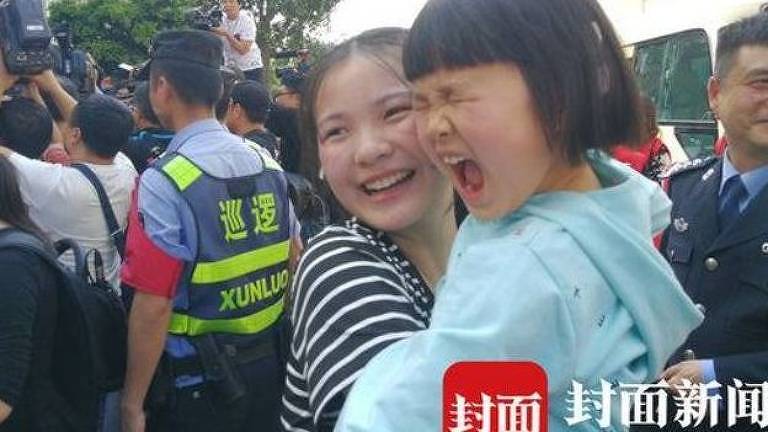 Sorrindo, Kang Ying abraça sua sobrinha, que franze os olhos e abre a boca; ao fundo, policiais e jornalistas formam uma rodinha