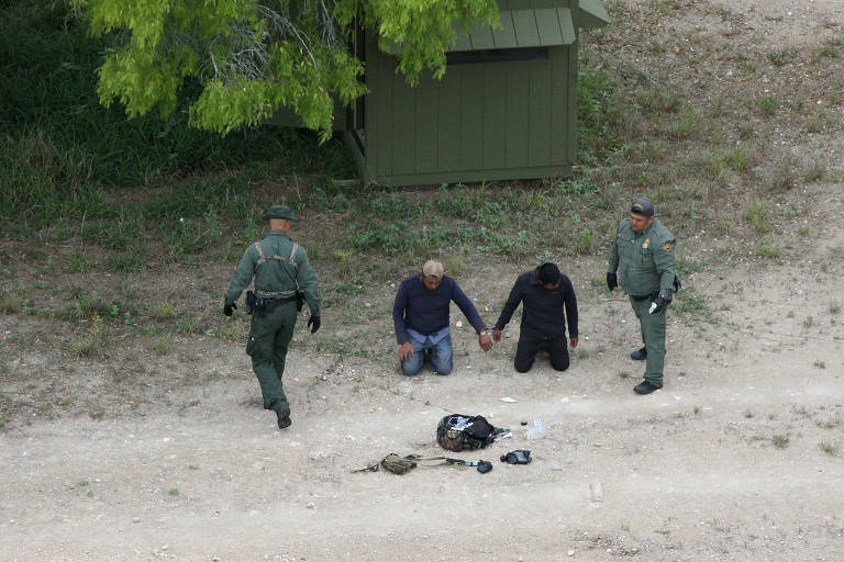 De roupa camuflada, dois agentes de fronteira circundam dois homens imigrantes que aparecem de mãos dadas em uma área ao lado de uma árvore; à frente dos imigrantes, uma mochila e outros pertences
