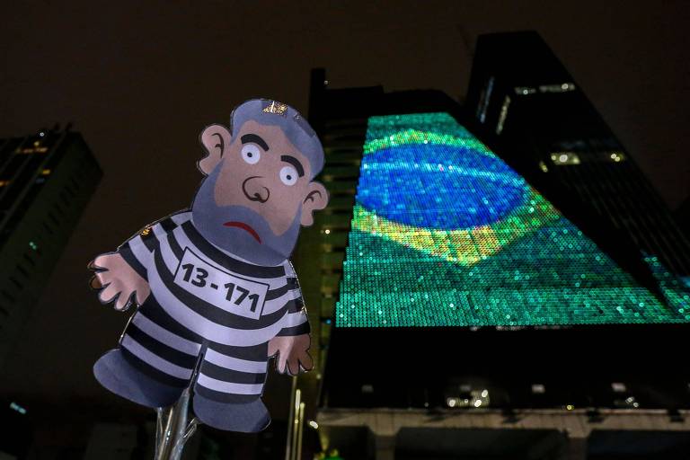 Boneco de Lula vestido de presidiário de papel aparece do lado do prédio da Fiesp, em São Paulo, em cuja frente há uma projeção da bandeira do Brasil