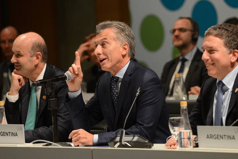 Macri aparece com o dedo indicador esquerdo enquanto fala durante evento; à sua direita, Nicolás Dujovne ri e, à esquerda, Federico Sturzenegger apoia seu queixo em sua mão esquerda