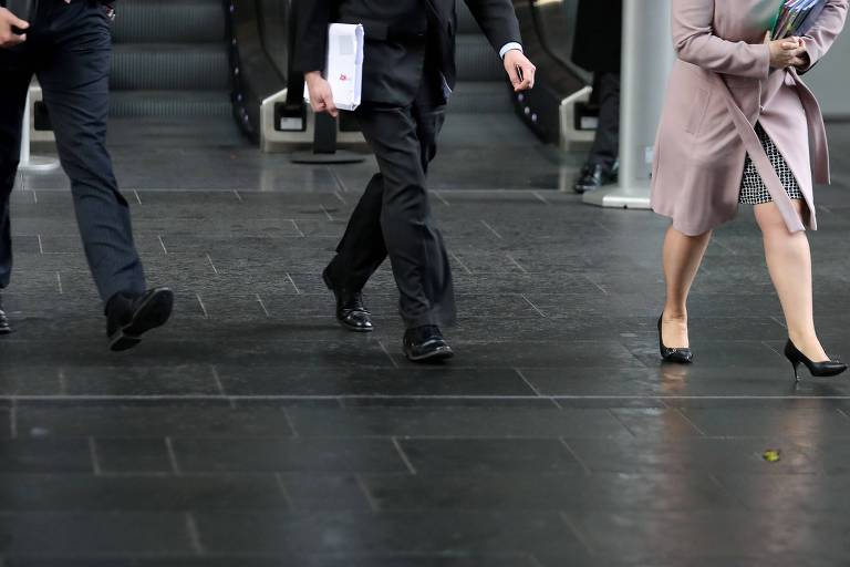 Pernas de mulher que veste saia e salto alto caminham à frente de pernas de dois homens que vestem terno escuro