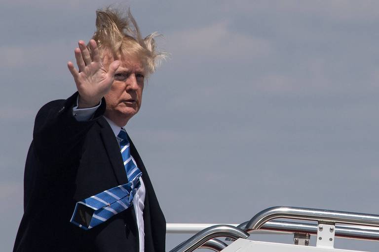Donald Trump acena com a mão do alto da escada de acesso ao Air Force One (não aparece na imagem); seu cabelo voa para o alto com o vento