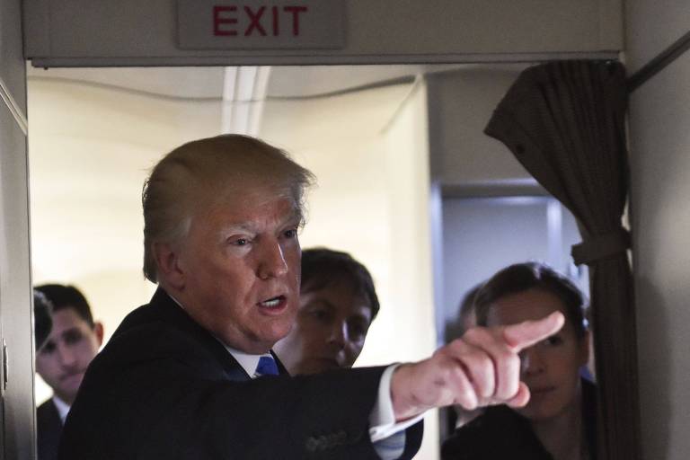 Trump estica o braço esquerdo e aponta para um repórter (não aparece na imagem) no corredor do Air Force One; ao fundo, três assessores observam presidente