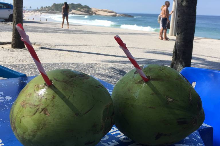 Dois cocos em cima de uma mesa com canudos enfiados neles. Ao fundo, a praia de Ipanema, no Rio de Janeiro
