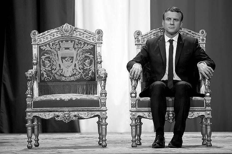 O presidente francês, Emmanuel Macron, está sentado em uma cadeira. À sua esquerda, há outra cadeira igual, mas vazia.