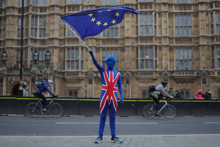 Uma pessoa pintada de azul e usando uma túnica com a estampa da bandeira do Reino Unido agita uma bandeira da União Europeia na calçada diante do prédio do Parlamento, em Londres