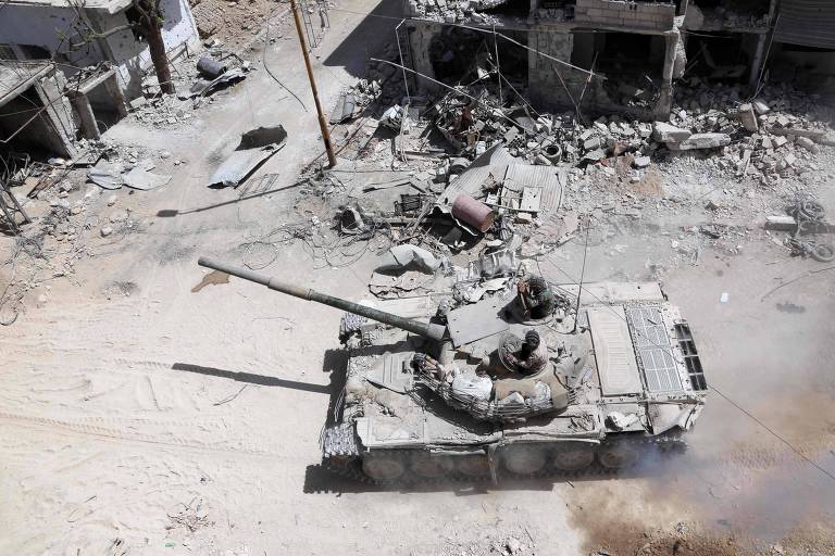 Dois soldados aparecem dentro de um tanque de guerra em uma área com construções destruídas; veículo blindado é visto de cima